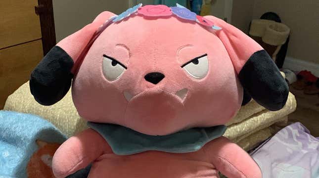 A Snubbull plush is seen laying on a bed and wearing a toy flower crown.